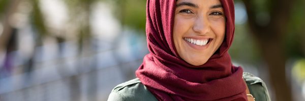 Portrait of young muslim woman wearing hijab head scarf in city while looking at camera. Closeup face of cheerful woman covered with headscarf smiling outdoor. Casual islamic girl at park.